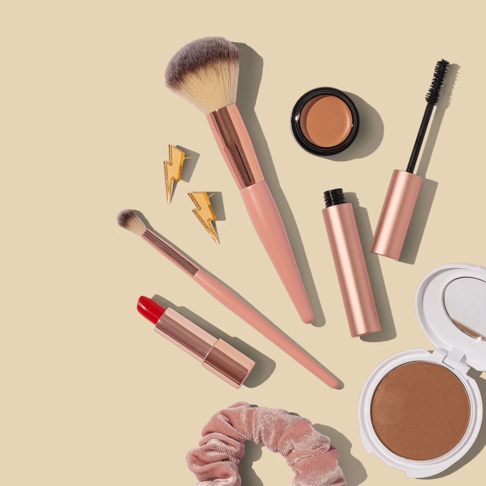 Photographie d’ambiance : pinceaux, mascara et rouge à lèvres sur fond beige.