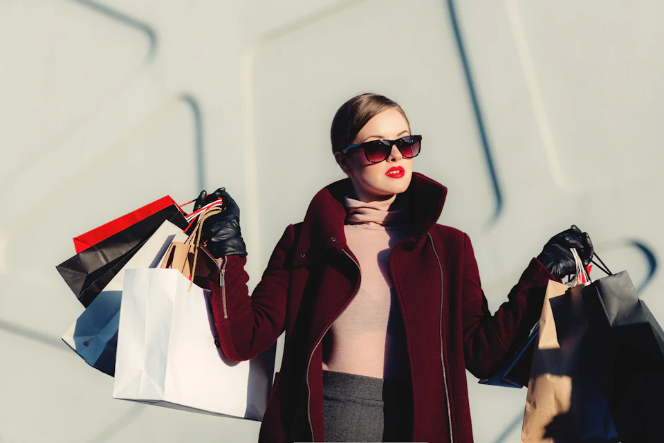 Photographie d’ambiance : une femme en manteau bordeaux et lunettes noires, des sacs à la main.
